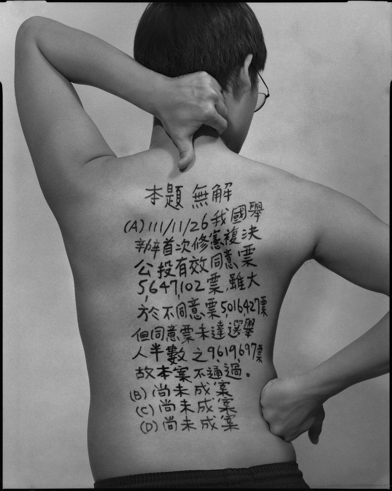 《答案》系列：黑白肖像，背面軀幹寫滿〈公民與社會，2029〉考題手寫解答，雙手於頸後或背後交扣。