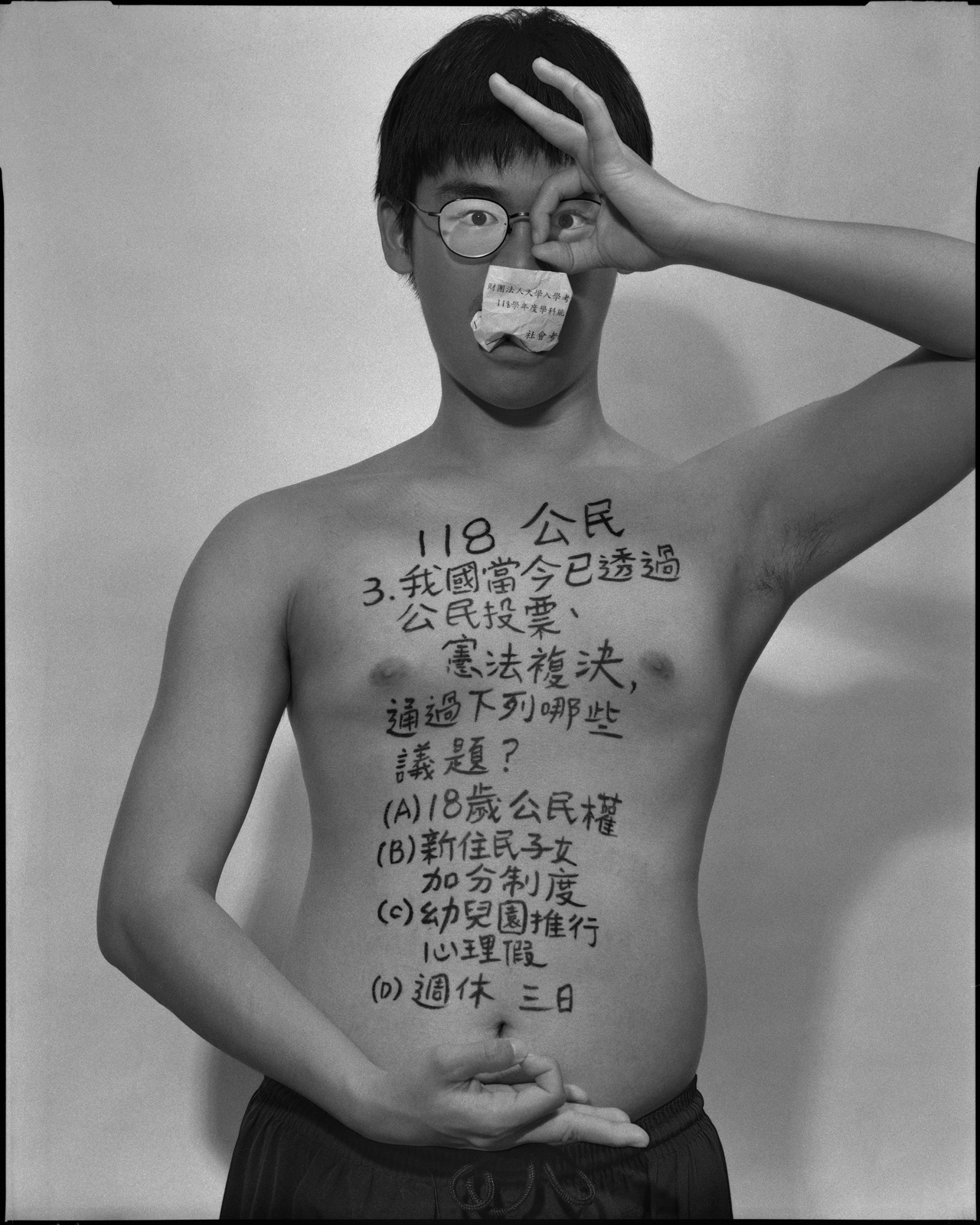 《答案》系列：黑白肖像，正面軀幹寫滿〈公民與社會，2029〉考題手寫文字，口中塞著紙捲，雙手於臉部前交疊。