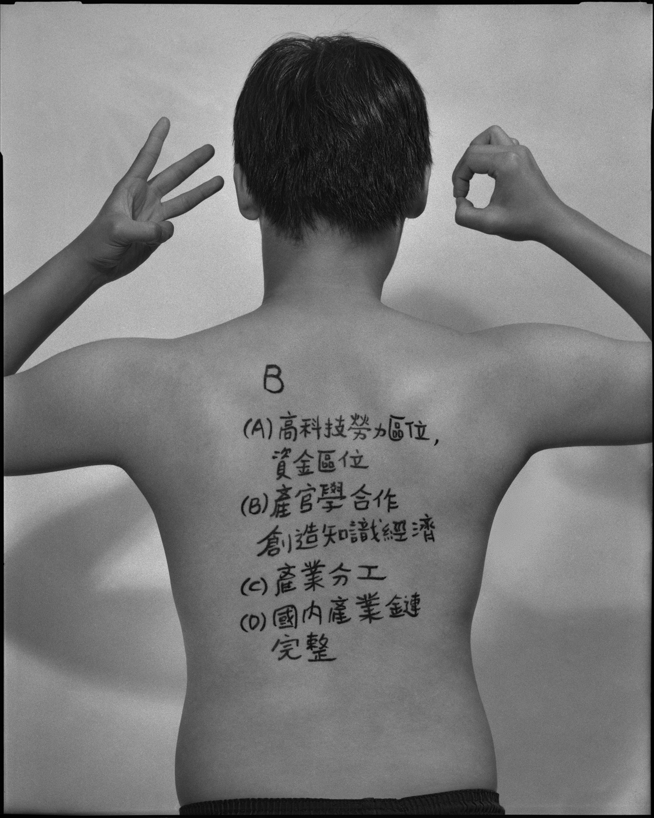 《答案》系列：黑白肖像，背面軀幹寫滿〈地理，2023〉考題手寫解答，雙手於頸後或背後交扣。