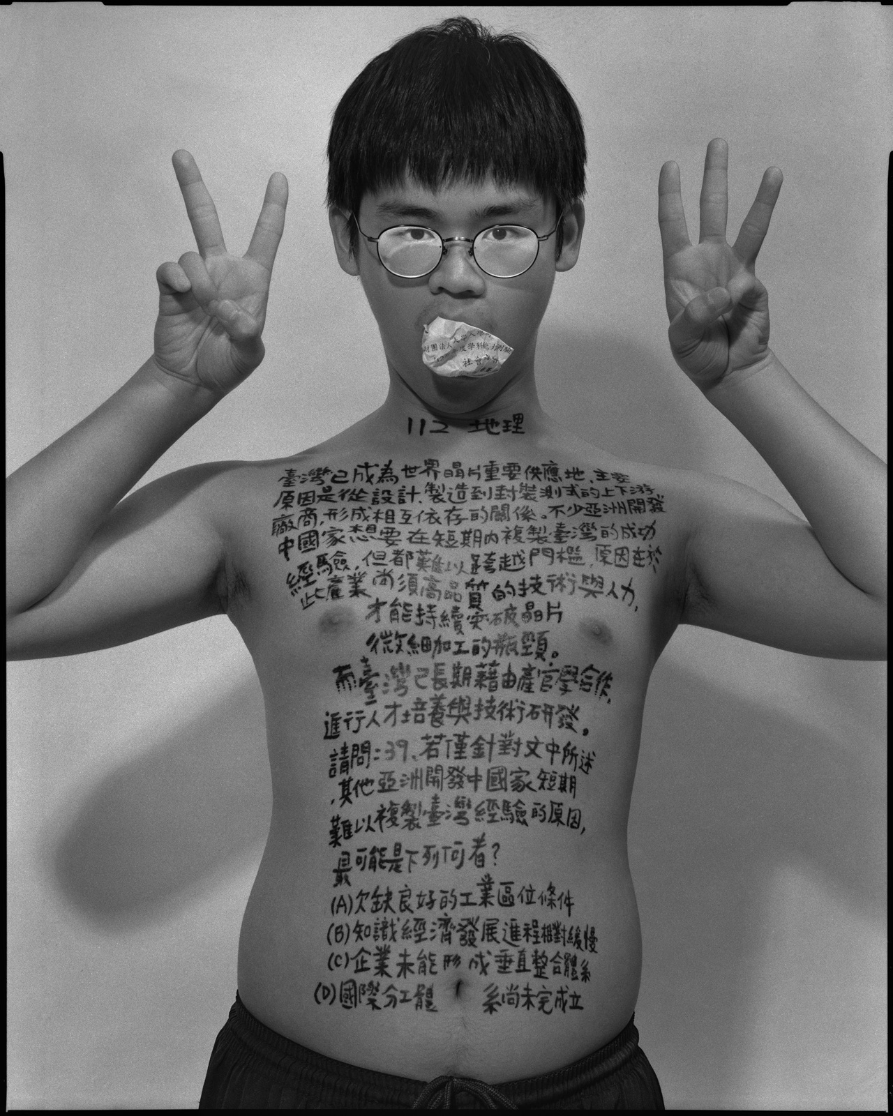 《答案》系列：黑白肖像，正面軀幹寫滿〈地理，2023〉考題手寫文字，口中塞著紙捲，雙手於臉部前交疊。
