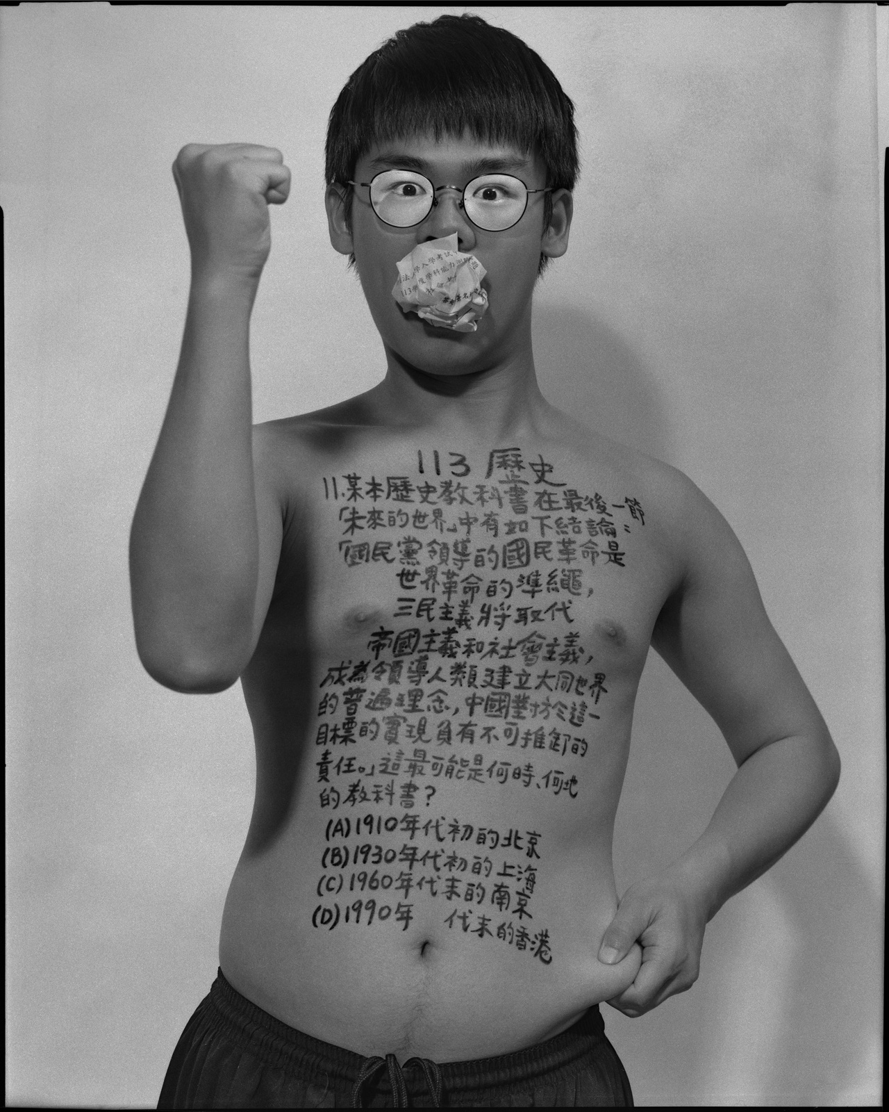 《答案》系列：黑白肖像，正面軀幹寫滿〈歷史，2024〉考題手寫文字，口中塞著紙捲，雙手於臉部前交疊。
