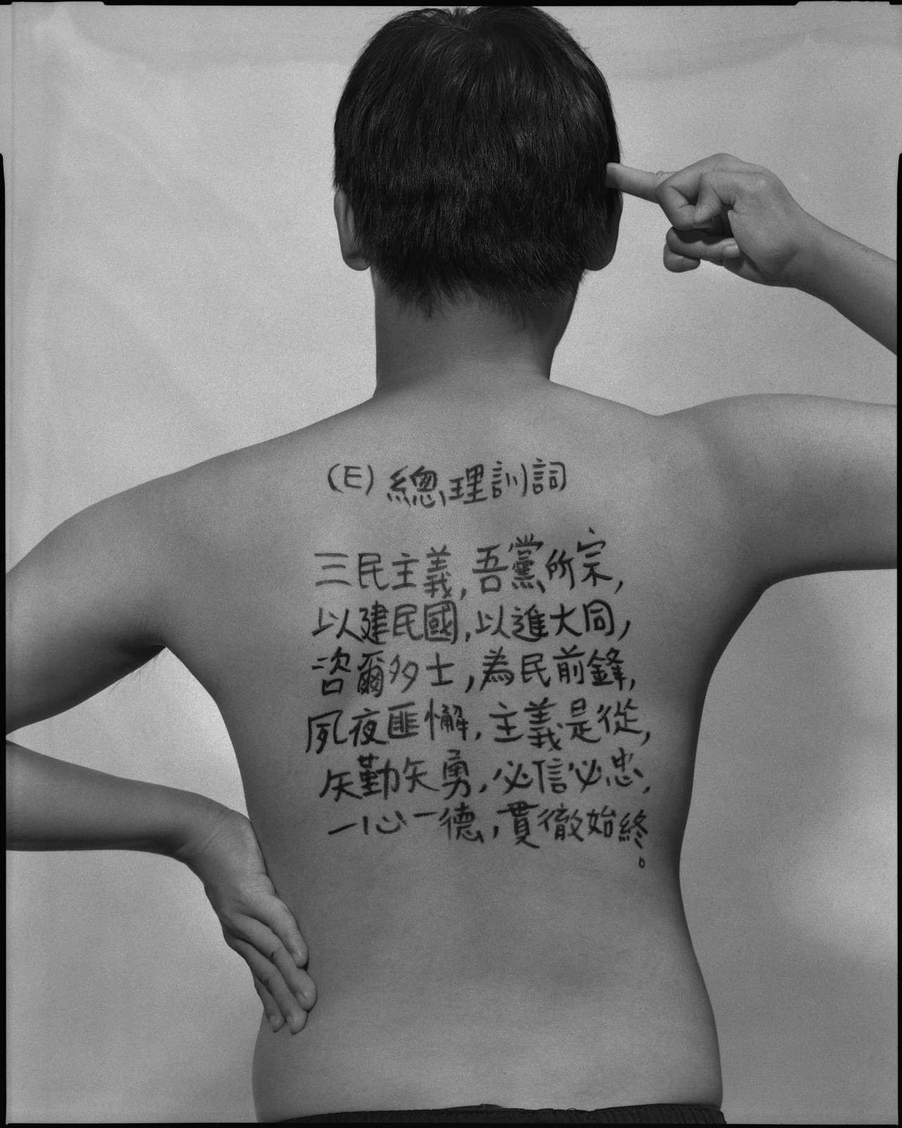 《答案》系列：黑白肖像，背面軀幹寫滿〈國防，2029〉考題手寫解答，雙手於頸後或背後交扣。