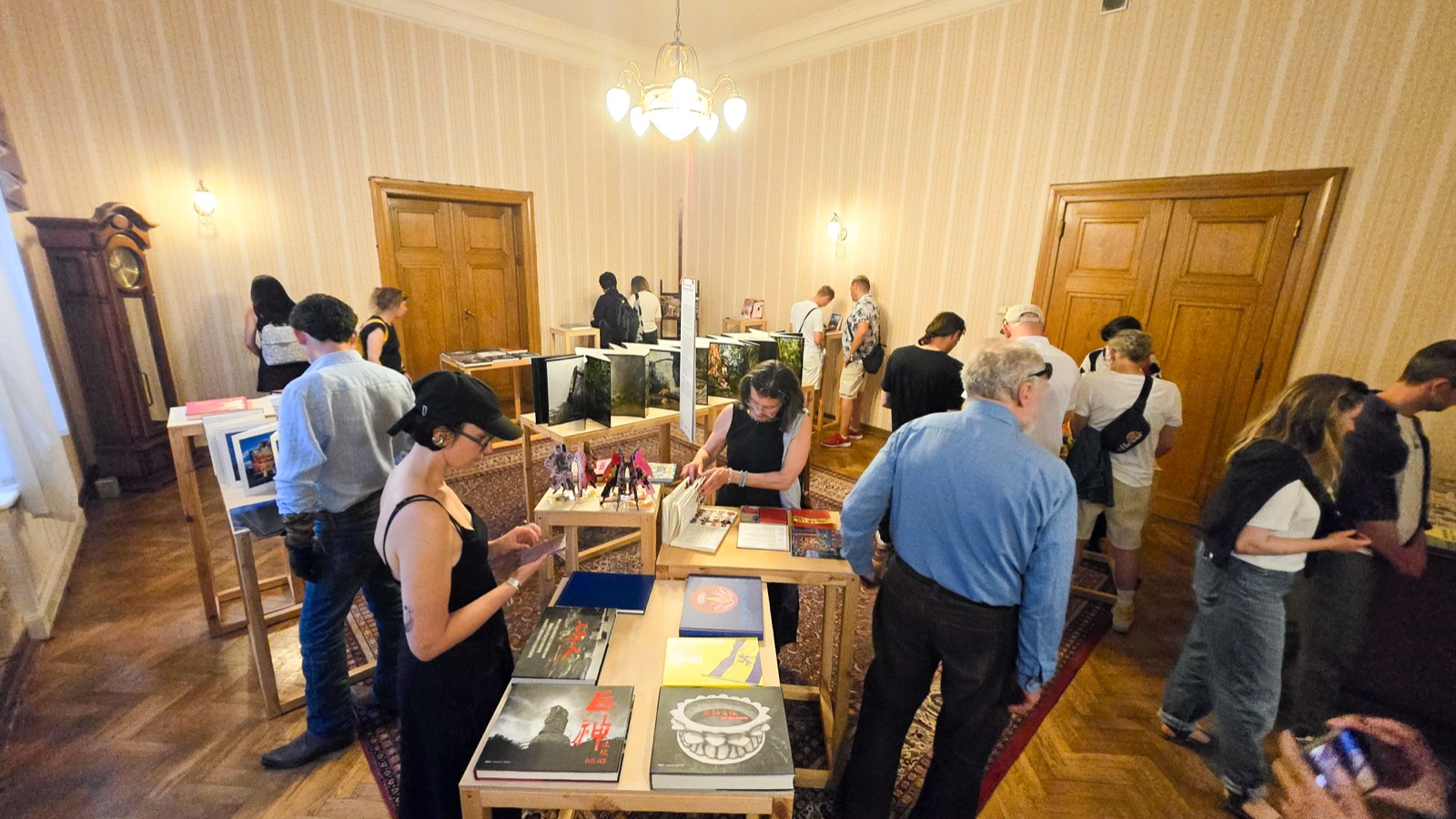 《複眼之島》羅茲展場：觀眾於古典展間圍著木質展桌翻閱臺灣藝術家書籍，上方吊燈、旁有落地鐘。