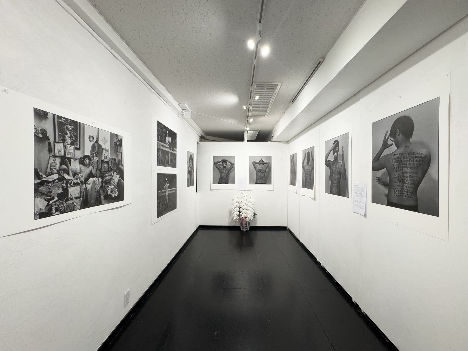 展場長廊一景：兩側牆面掛滿《答案》系列黑白輸出，走道盡頭擺放白色蘭花。